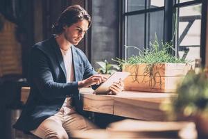 Working just from cafe. Confident young man in smart casual wear working on digital tablet while sitting near window in cafe photo