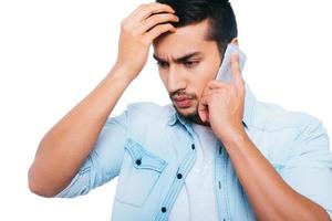 malas noticias. joven indio frustrado hablando por teléfono móvil y tomándose la mano en el cabello mientras se enfrenta a fondo blanco foto