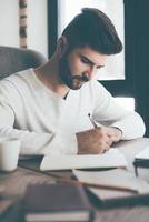 haciendo algunas notas. un joven seguro de sí mismo escribiendo algo en un cuaderno mientras se sienta en su lugar de trabajo en la oficina foto