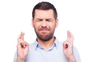 Waiting for special moment. Portrait of young man in shirt keeping fingers crossed and eyes closed while standing against white background photo