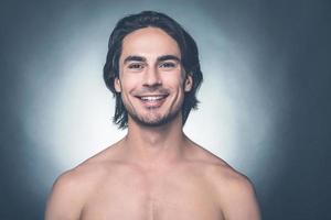 Looking good and feeling great. Portrait of young shirtless man looking at camera and smiling while standing against grey background photo