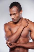 Feeling pain in elbow. Young muscular African man touching his elbow while standing against grey background