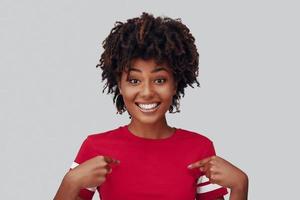 Attractive young African woman looking at camera with smile and pointing at herself while standing against grey background photo