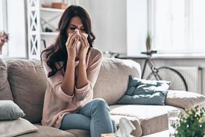 Cold and flu. Sick young woman blowing the nose using tissue paper while sitting on the sofa at home photo