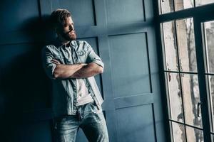 Thinking about... Handsome young bearded man in light jeans shirt keeping arms crossed and looking away while standing near the window photo