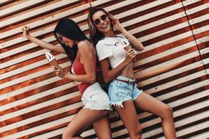 disfrutando de un fin de semana loco. dos mujeres jóvenes atractivas sonriendo y sosteniendo un helado mientras están de pie contra la pared de madera al aire libre foto