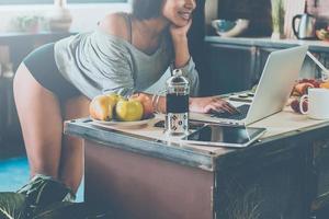 Surfing web from kitchen. Close-up of attractive young African woman leaning at the kitchen desk and looking at laptop with smile photo