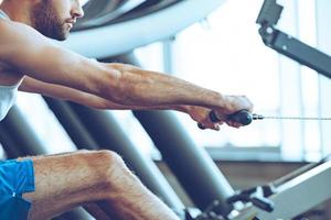 Row and pull. Side view close-up part of young man in sportswear doing rowing in front of window at gym photo