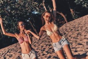 dos atractivas mujeres jóvenes en pantalones cortos y trajes de baño sonriendo mientras corren en la playa foto