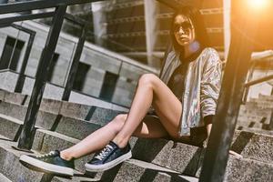 Enjoying nice day. Beautiful young Asian woman in casual wear looking at camera while sitting on the stairs outdoors photo