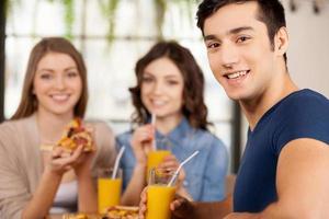 Friends eating pizza. Three cheerful young people eating pizza at the restaurant while man looking over shoulder and smiling at camera photo