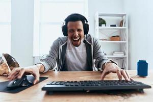 despreocupado y feliz. hombre joven guapo con ropa informal mirando a la cámara y sonriendo mientras juega juegos de computadora en casa foto