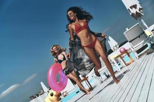 verano divertido. Longitud total de dos hermosas mujeres jóvenes en bikini corriendo juntas cerca de la piscina mientras una de ellas lleva un anillo inflable foto