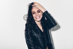 Feeling carefree. Beautiful young smiling woman in unbuttoned leather jacket holding hand in hair and keeping eyes closed while standing against grey background photo