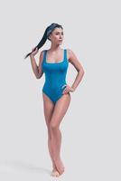 Elegance and beauty. Full length studio shot of attractive young woman in blue swimsuit touching her hair and looking away while standing against grey background photo