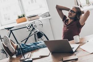 Time to relax. Top view of handsome young man with long hair holding hands behind head and looking relaxed while sitting at his working place in office photo