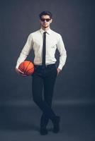 listo para jugar toda la longitud de un joven apuesto y bien vestido con gafas de sol sosteniendo una pelota de baloncesto y mirando la cámara mientras se enfrenta a un fondo gris foto