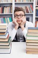 nerd soñando. joven aburrido con camisa y corbata de moño sentado en la mesa de la biblioteca y sosteniendo la cabeza en las manos mientras dos pilas de libros están en primer plano foto