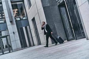 charla rápida de negocios. toda la longitud de un joven con traje completo hablando por teléfono y sonriendo mientras camina al aire libre foto