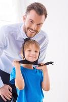 Playing with necktie. Cheerful little boy playing with necktie of his father standing near him and smiling at camera photo