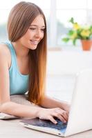 Surfing the net at home. Beautiful young teenage girl using computer while lying down on the floor at her apartment photo