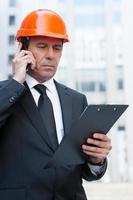 Busy contractor. Confident mature man in formalwear and hardhat talking on the mobile phone and looking at clipboard while standing outdoors photo