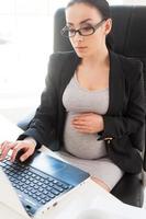 Pregnant businesswoman. Beautiful pregnant businesswoman working on laptop while sitting at her working place in office photo