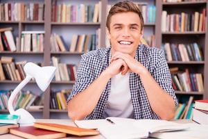 Enjoying his time in library. Handsome young man leaning his face on hands and smiling while sitting at the desk in library photo