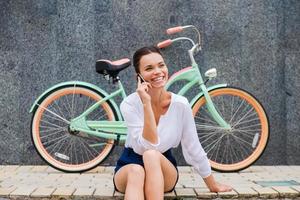 buena charla con un amigo. atractiva joven mujer sonriente hablando por teléfono móvil mientras se sienta en la carretera cerca de su bicicleta vintage foto