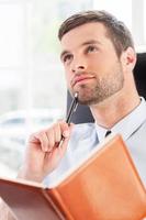 Business dreams. Thoughtful young man in shirt and tie holding note pad and touching chin with pen while sitting at his working place photo