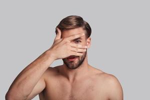 I want to know everything. Handsome young man covering eye with hand and peeking while standing against grey background photo