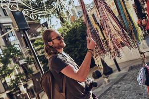 What to choose Handsome young man in casual clothing choosing necklace and smiling while standing near the market stall outdoors photo