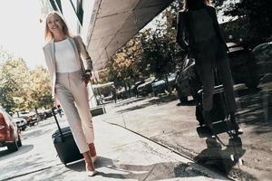 joven profesional de negocios. Longitud total de mujer joven en traje tirando de equipaje y sonriendo mientras camina al aire libre foto