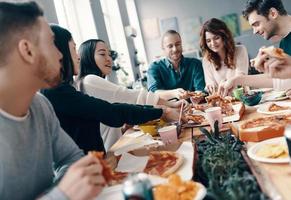 tener hambre. grupo de jóvenes con ropa informal recogiendo pizza y sonriendo mientras cenan en el interior foto