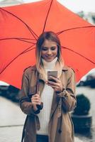 Quick message on the go. Attractive young smiling woman carrying umbrella and looking at her mobile phone while walking by the street photo