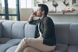 Handsome young man talking on smart phone and looking away while sitting on the sofa photo