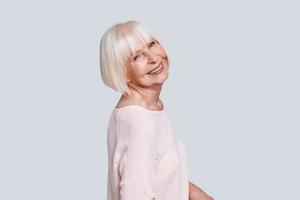 Looks perfect. Beautiful senior woman looking at camera and smiling while standing against grey background photo