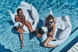 No better way to spend summer. Top view of two beautiful young women in bikini looking away and smiling while floating on big inflatable swans photo