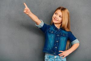 Take a look here Cheerful littlegirl pointing away and smiling while standing against grey background photo