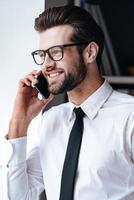 Great news Cheerful young handsome businessman in glasses talking on the mobile phone and smiling while standing in office photo