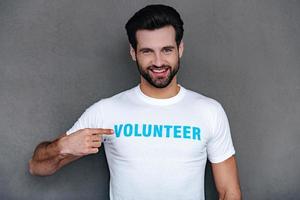 I can help you Confident young man pointing at volunteer title on his t-shirtand looking at camera with smile while standing against grey background photo