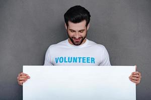 por favor ayude a un joven confiado en la comunidad con una camiseta voluntaria sosteniendo una pizarra blanca y mirándola con una sonrisa mientras se enfrenta a un fondo gris foto