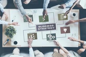 Discussing company strategy. Top view of business people pointing large paper with conceptual business icons while sitting together at the desk photo