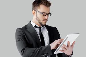 Confident in his gadget. Cheerful young man in formalwear and glasses working on digital tablet while standing against grey background photo