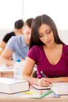 Confident student. Beautiful female student writing something in note pad while sitting in classroom with other students photo