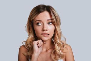 Asking herself. Confused young woman looking away and biting lip while standing against grey background photo