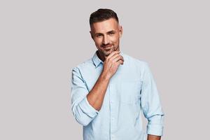 Positively charming. Handsome young man looking at camera and keeping hand on chin while standing against grey background photo