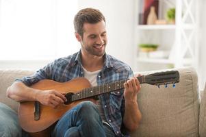 Composing his new song. Happy young man playing the guitar while sitting on sofa at home photo