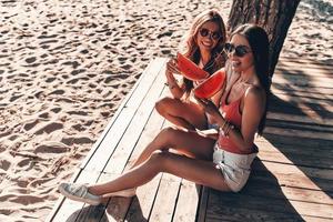 Let the relaxation begin. Top view of two attractive young women smiling and eating watermelon while sitting on the beach photo
