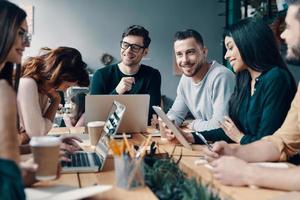 Putting ideas into something real. Group of young modern people in smart casual wear discussing something and smiling while working in the creative office photo